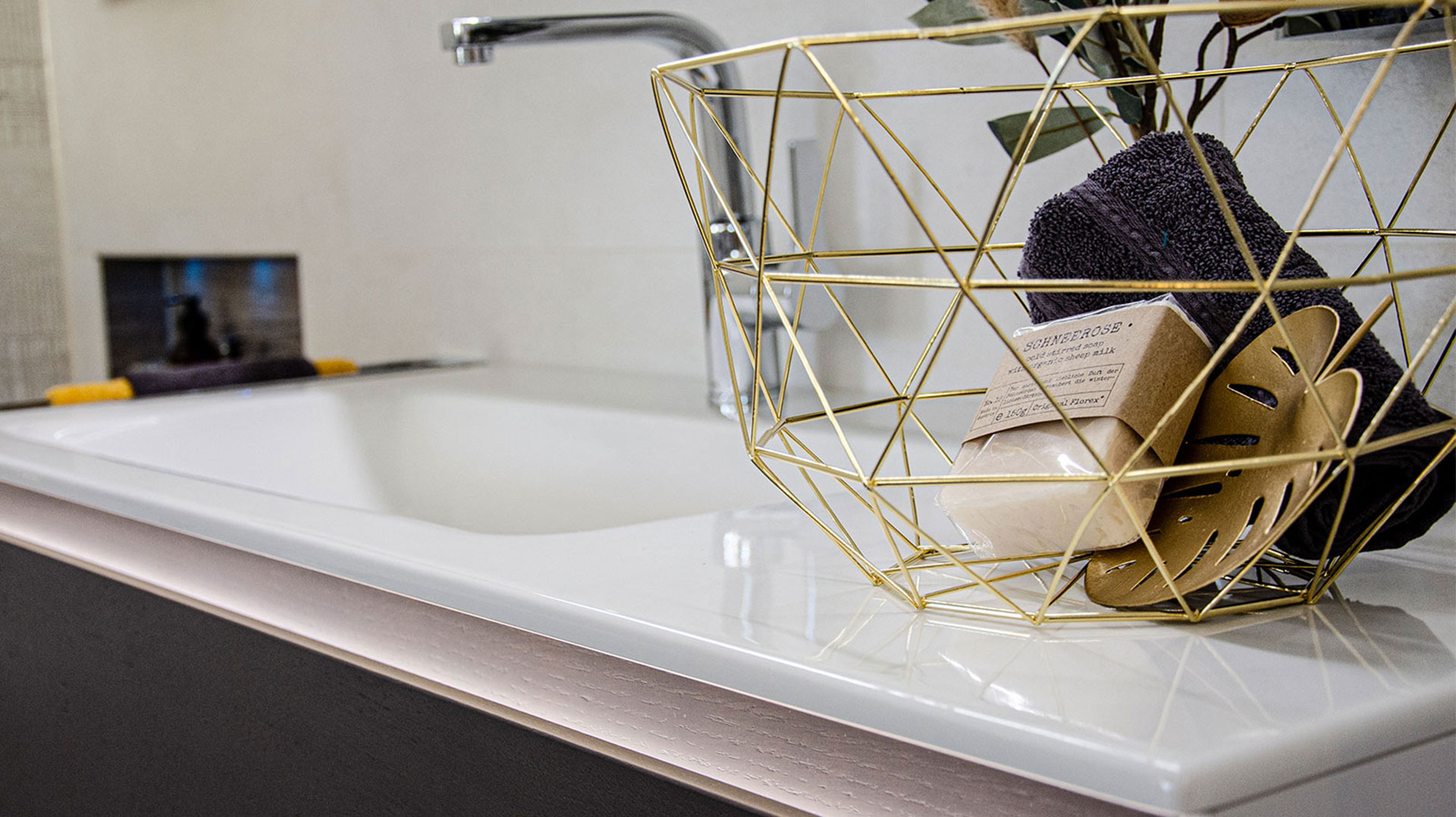 Detailansicht einer weißen Badewanne mit Chromarmatur. Auf dem Wannenrand steht ein goldfarbener Drahtkorb mit Handtuch und Seife. Moderne, helle Badgestaltung.